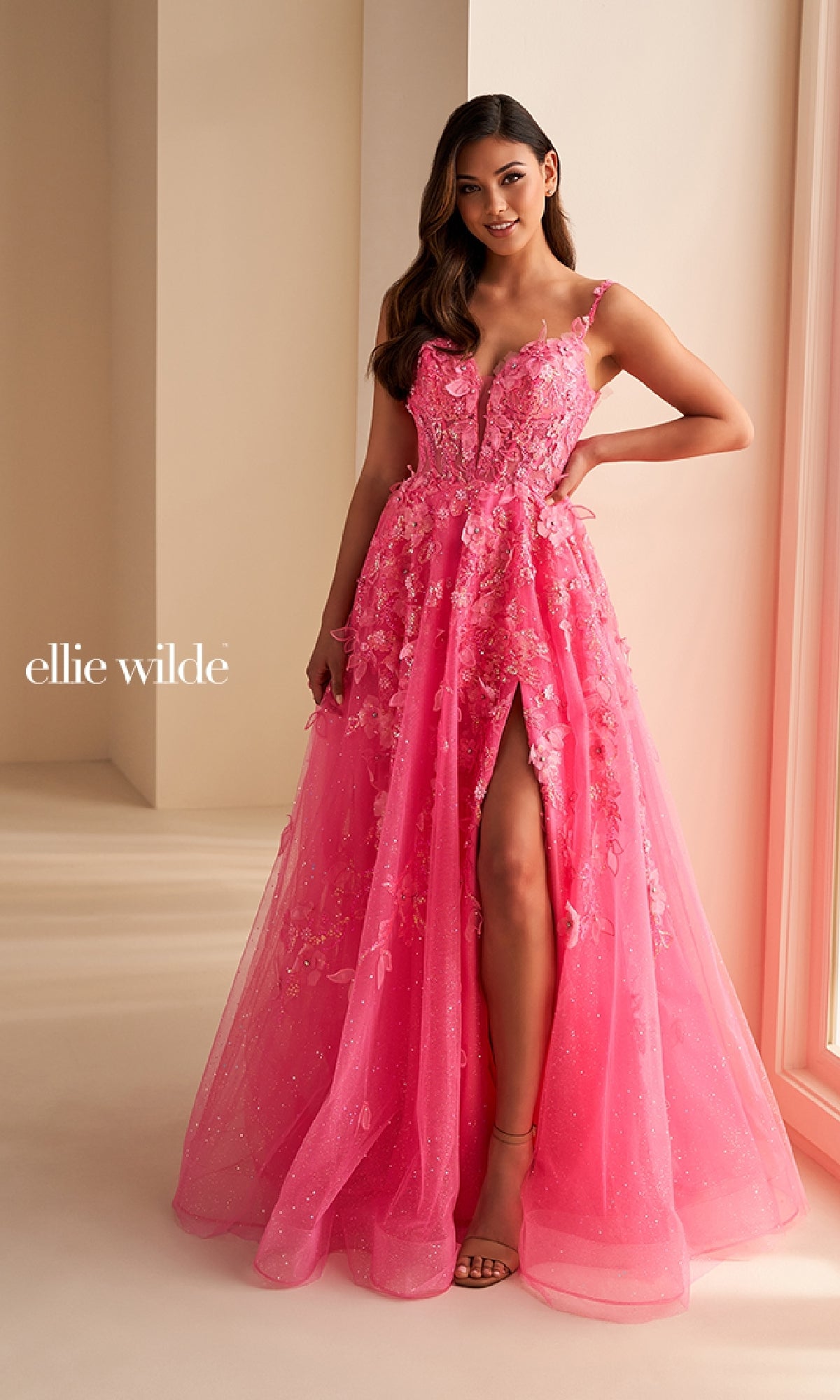 A woman poses by a window in the Ellie Wilde EW36208, a bright pink floor-length prom dress featuring glitter tulle, 3D flowers, a slit, and spaghetti straps.